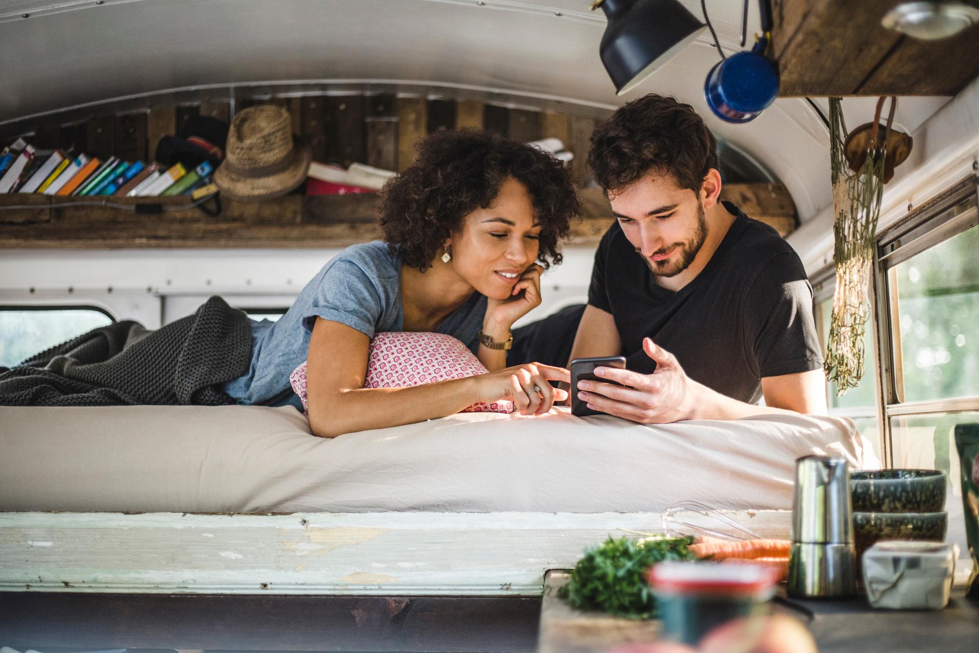 Junges Paar liegend im Campervan, beide schauen auf ein Smartphone