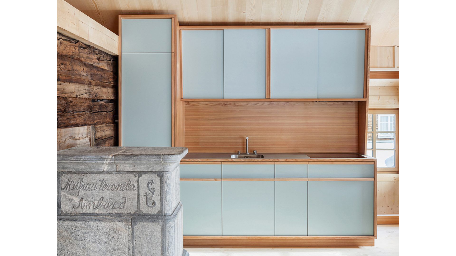 En avant-plan, on peut voir l'un des fourneaux en pierre ollaire rénovés. La nouvelle cuisine est en mélèze, et l'aménagement intérieur entièrement réalisé en bois de sapin, crée un contraste avec les murs sombres en madrier. Les baguettes de couverture confèrent aux nouvelles surfaces en bois profondeur et un caractère multidimensionnel. (Photo: Markus Käch, Emmenbrücke)