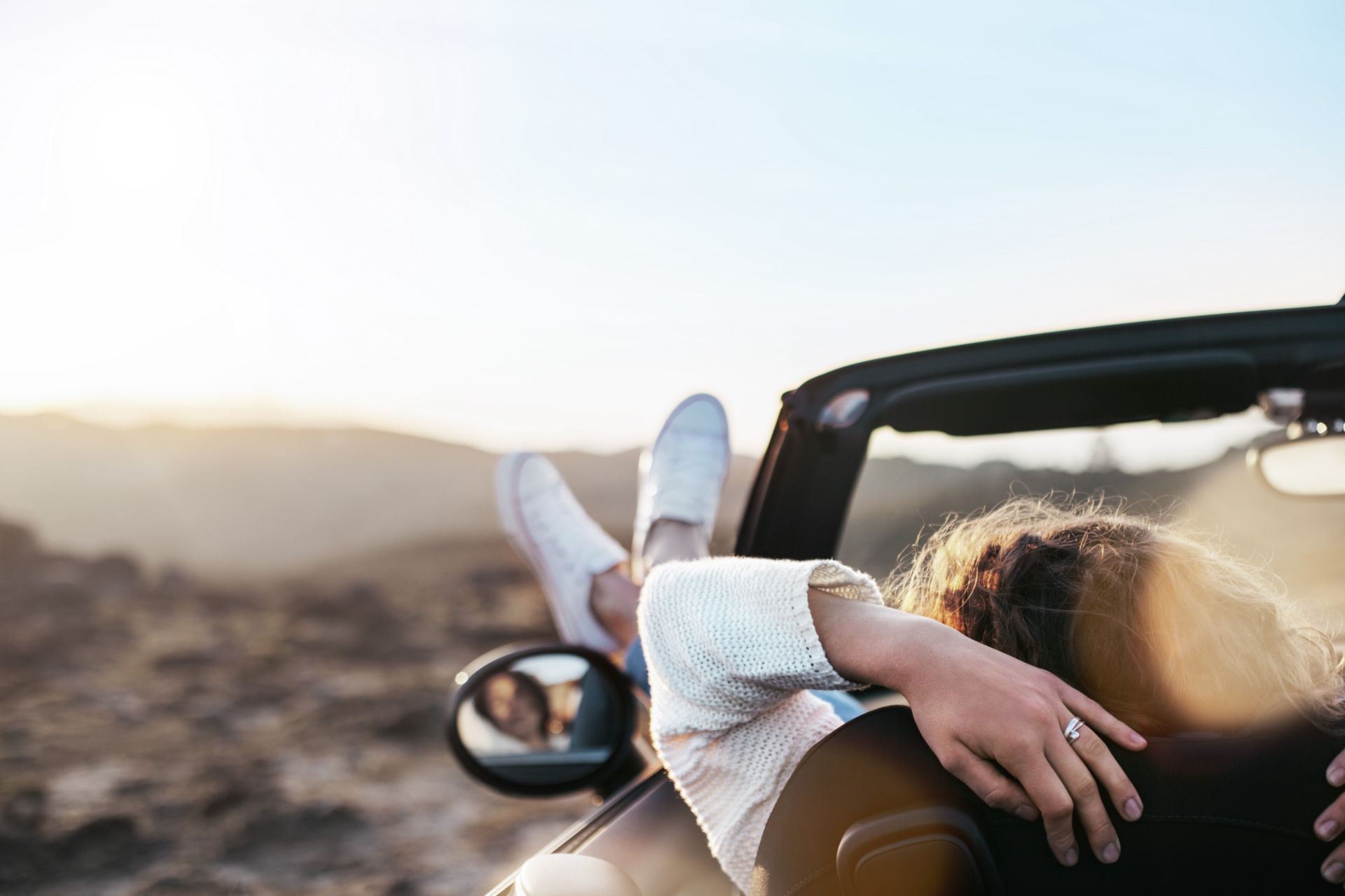 Eine Person mit langen Haaren und weissem Pullover lehtn entspannt aus einem Cabrio-Auto mit hochgelegten Beinen und weissen Schuhen, während die Sonne im Hintergrund untergeht.