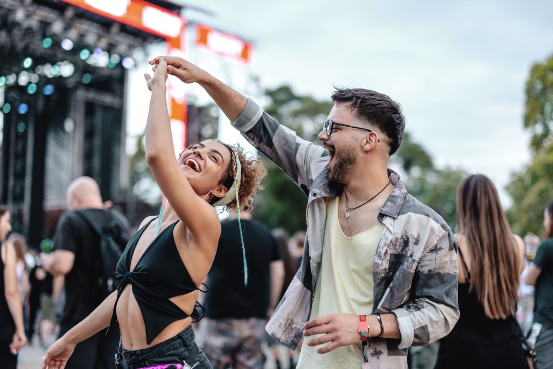 Due persone ballano all'aperto durante un festival o concerto. La persona a sinistra indossa una maglietta nera e ha i capelli ricci legati in alto. La persona a destra indossa una giacca mimetica sopra una maglietta chiara. Sullo sfondo si vedono altre persone e luci del palco.