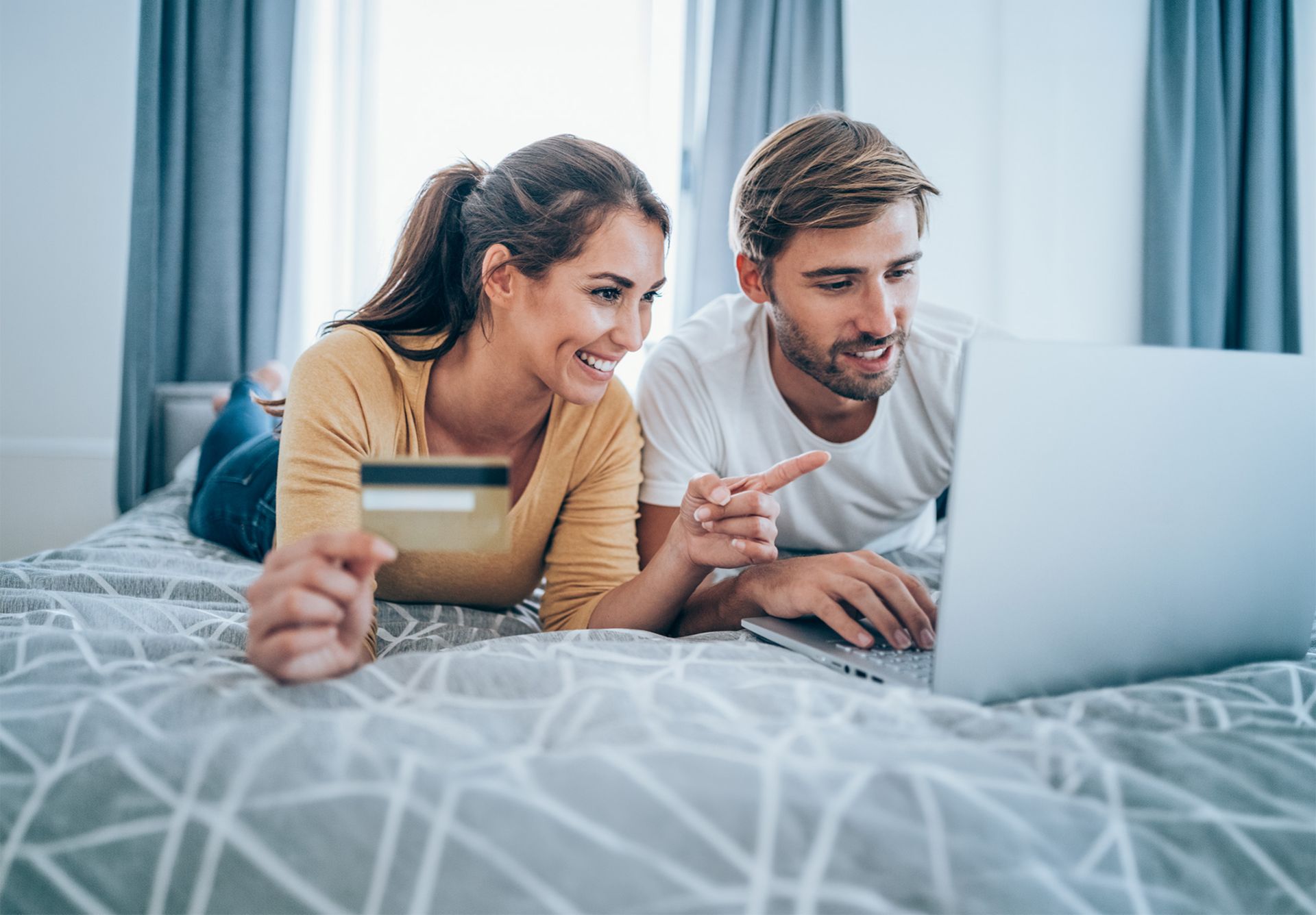 Une femme et un homme visiblement joyeux, allongés sur le lit, réservant leurs vacances avec une carte de crédit devant un ordinateur portable