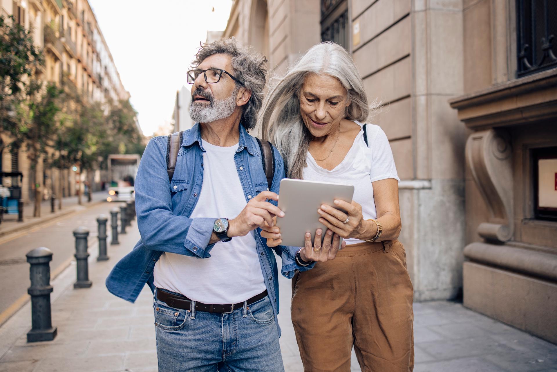 Älteres Touristenpaar, Frau und Mann, laufen durch eine Stadt und halten ein Tablet in der Hand.