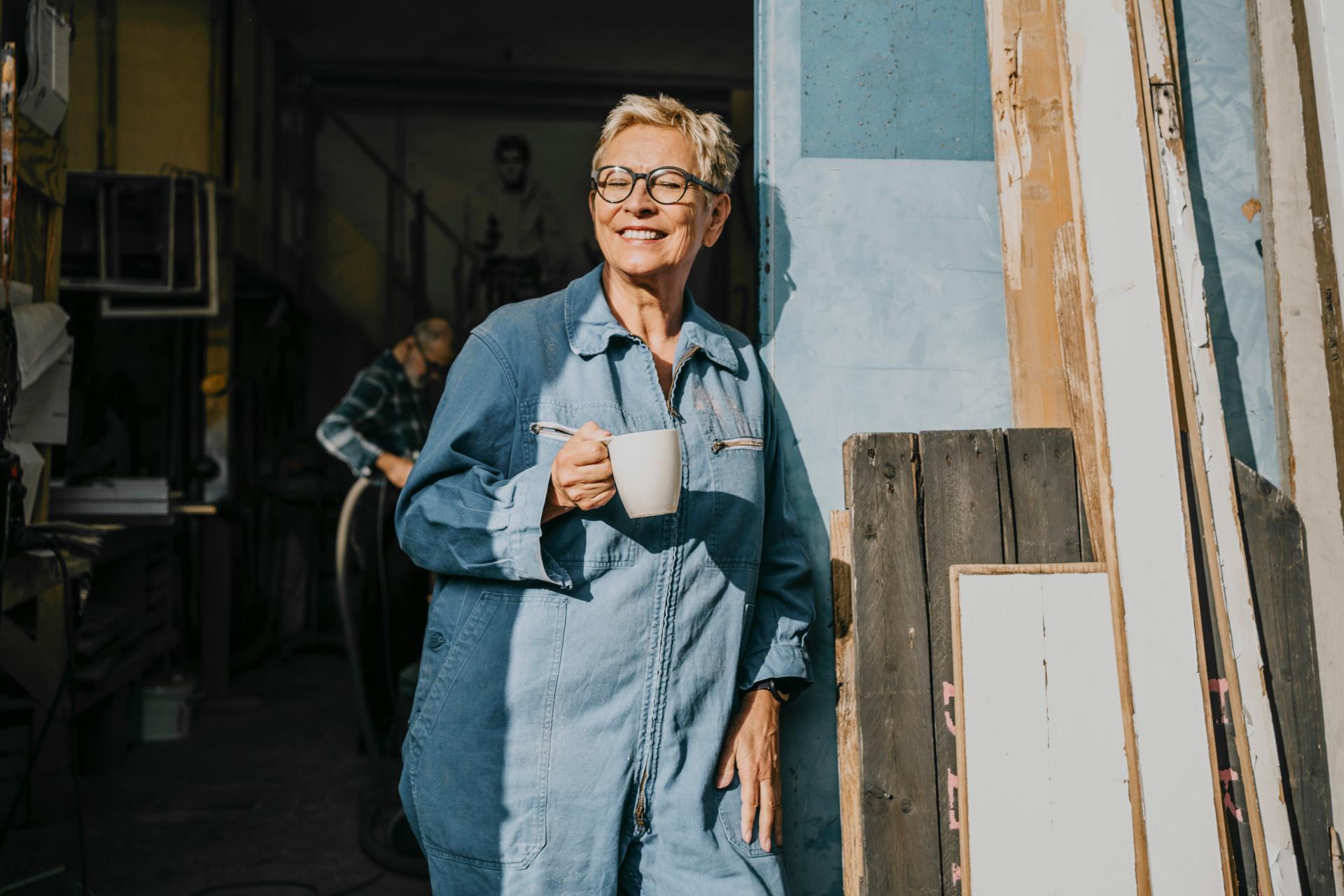 Ältere Dame steht mit einer Tasse in der Hand vor einer Werkstatt